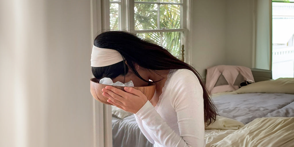 Woman in a white top and hair headband icing her face in a bowl, in a bright bedroom with a window in the background.