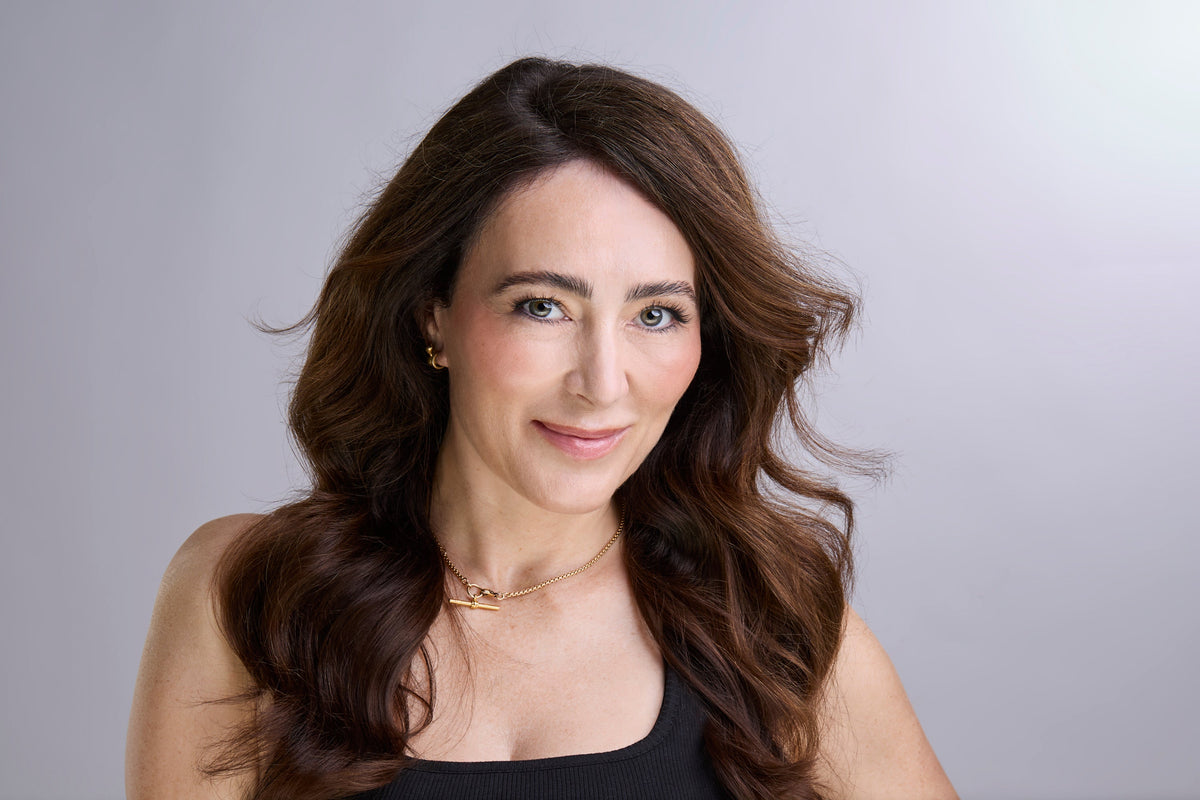 Headshot of Melanie Simon, founder of ZIIP Beauty, with wavy brown hair, wearing a black top against a soft grey backdrop.