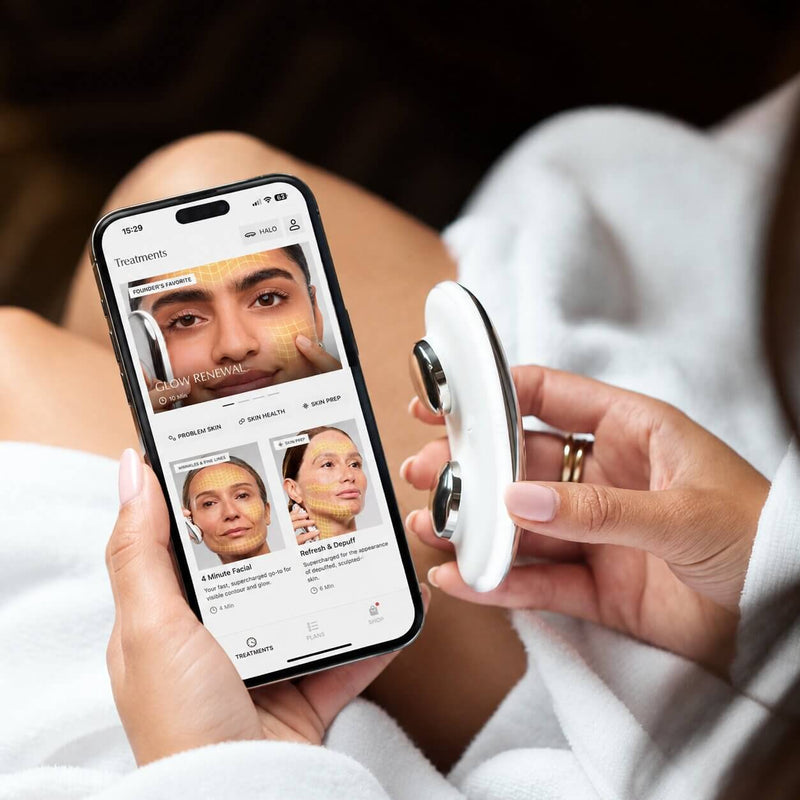 A person in a white robe holds a smartphone showing a skincare treatment app screen while holding a small white handheld device with black circular accents. The scene is cropped closely on the hands and lap, with a dark blurred background.